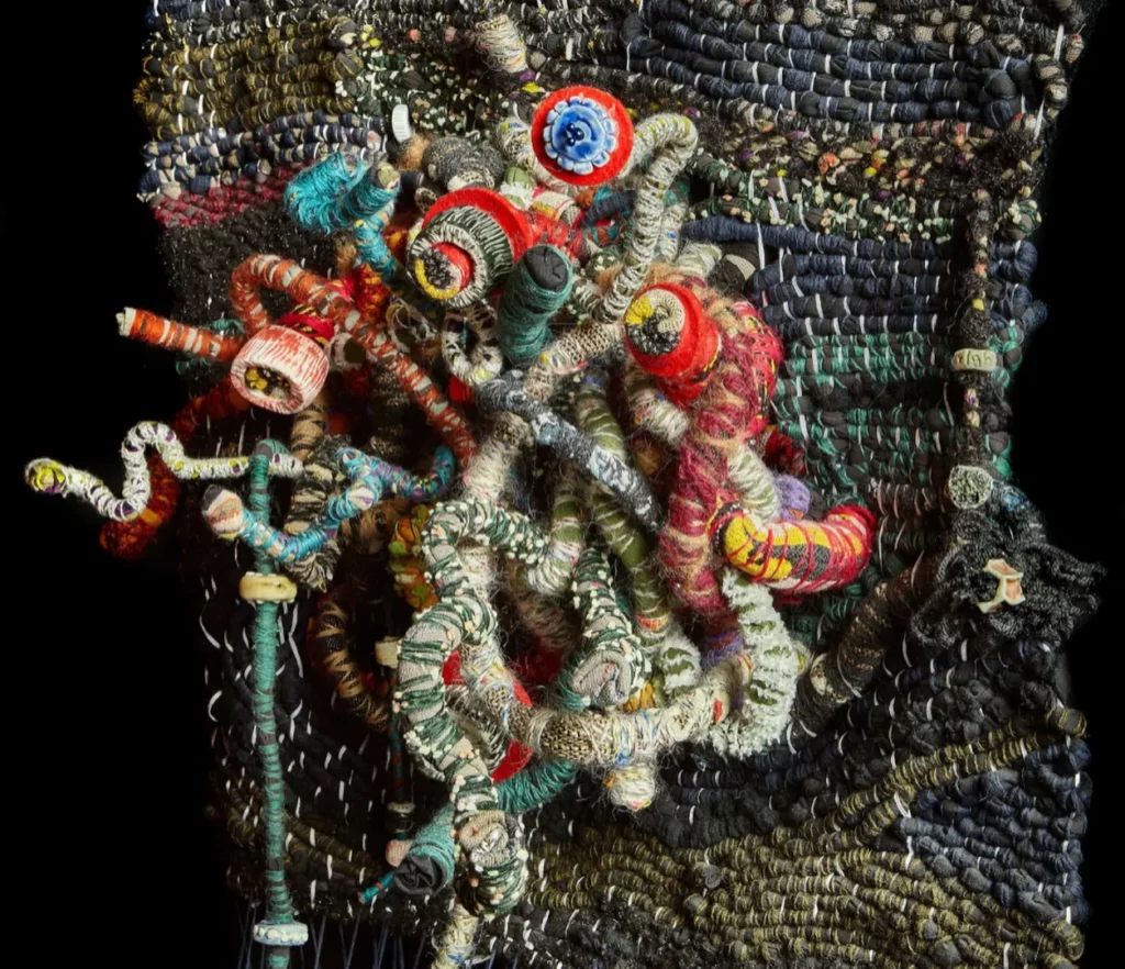 Enterlaced (Tal Waldman 2020) Crédit photo : Jonathan Goor
Dimensions : 100 x 120 x 20 cm et 50 cm de diamètre. Une installation composée d’une tapisserie tissée et d’une sculpture suspendue. Réalisée à l’aide de techniques de tissage et d’enroulement, à partir de laine, de coton, de métal souple et de textiles recyclés, cette œuvre s’inscrit dans le cadre du projet Enterlaced, réflexion sur une société entrelacée et son potentiel. Réalisée lors du premier confinement, elle symbolise le chaos et l’espace de transformation de cette période. Biennale AKA 2020 – Galerie HCE.